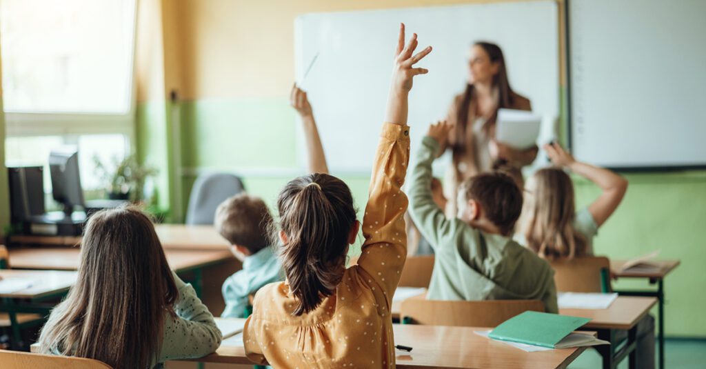 Students raising hands at a Jewish Day School while teacher asking them questions in classroom